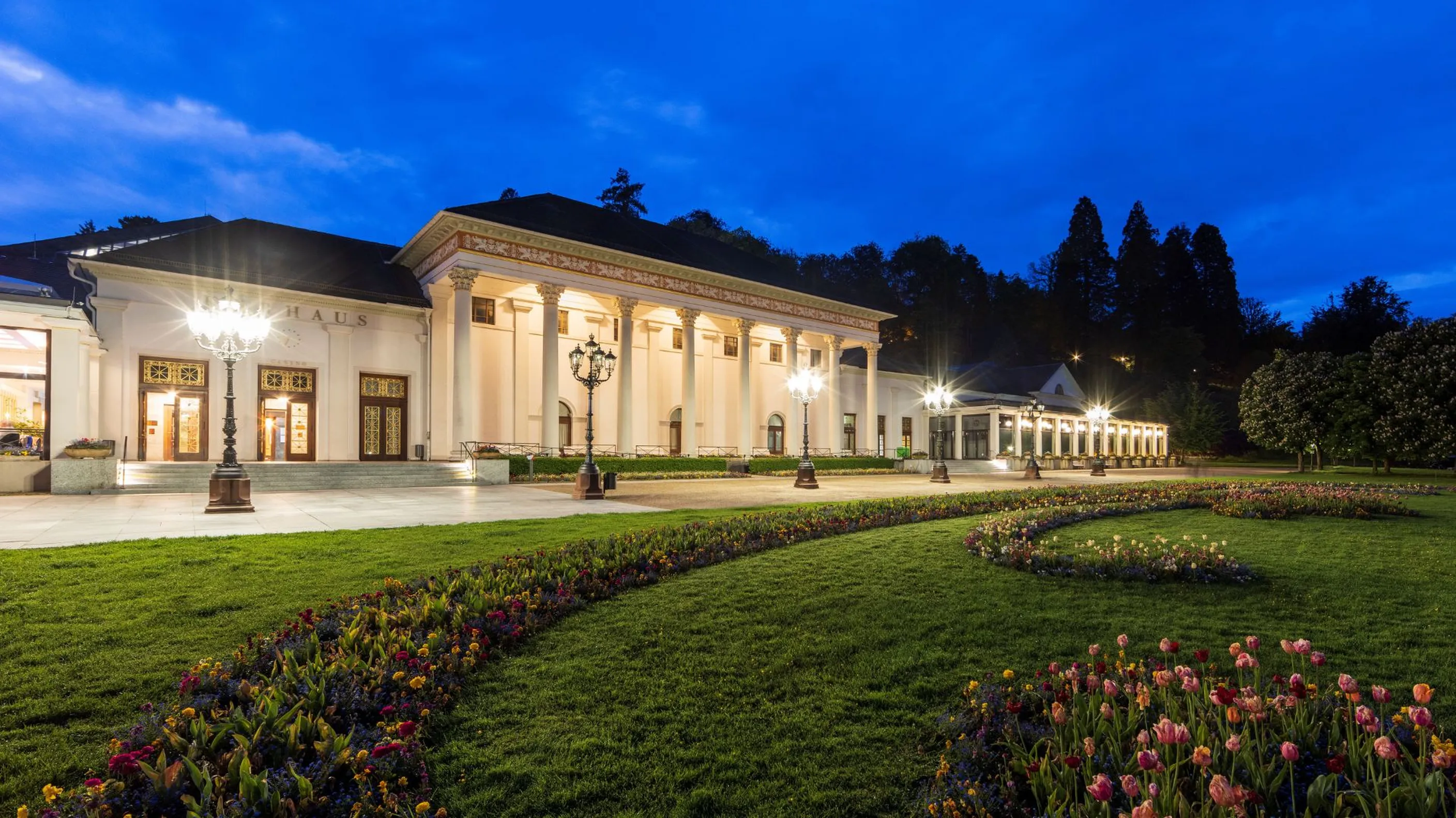 Beleuchtetes Kurhaus Baden-Baden bei Abenddämmerung mit dem Kurgarten und blühenden Beeten im Vordergrund.