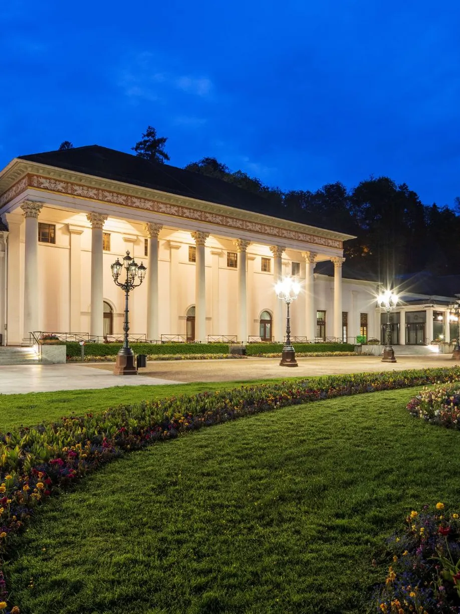 Beleuchtetes Kurhaus Baden-Baden bei Abenddämmerung mit dem Kurgarten und blühenden Beeten im Vordergrund.