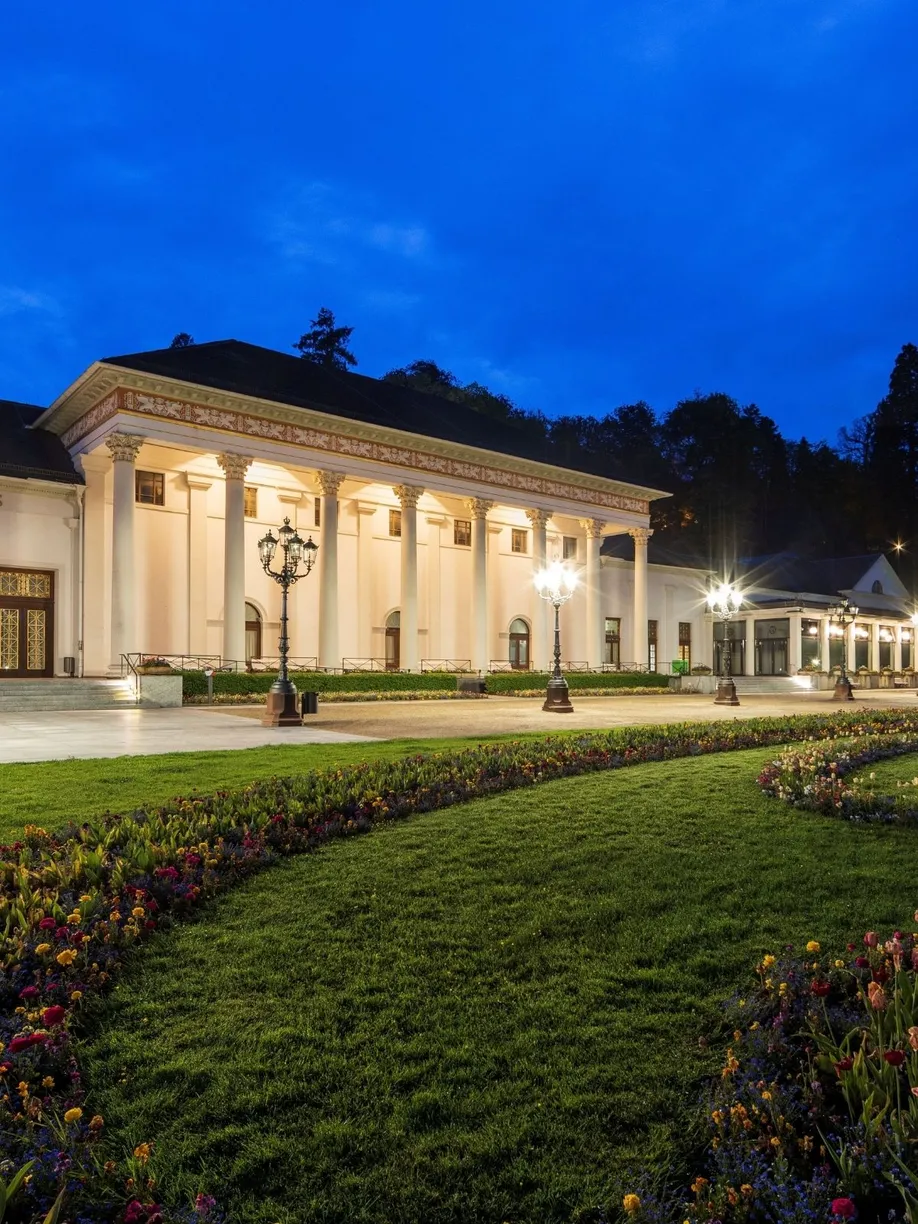 Beleuchtetes Kurhaus Baden-Baden bei Abenddämmerung mit dem Kurgarten und blühenden Beeten im Vordergrund.